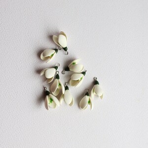 May include: A collection of small, white snowdrop flower charms with green accents and silver-colored metal loops. The charms are arranged on a white surface, showcasing their delicate, handcrafted details. These charms are ideal for jewelry making or craft projects.