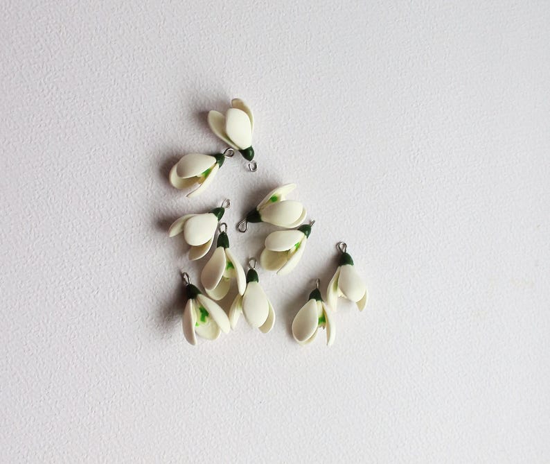 May include: A collection of small, white snowdrop flower charms with green accents and silver-colored metal loops. The charms are arranged on a white surface, showcasing their delicate, handcrafted details. These charms are ideal for jewelry making or craft projects.