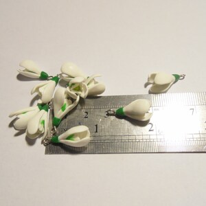 May include: Several small, white, handcrafted snowdrop flower charms with green stems and silver-colored jump rings. A metal ruler is next to the charms, showing measurements in inches.