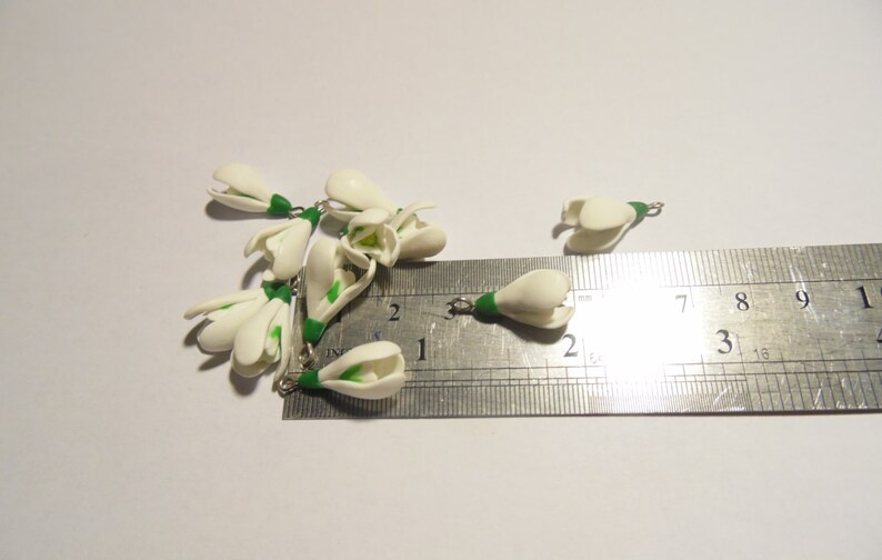 May include: Several small, white, handcrafted snowdrop flower charms with green stems and silver-colored jump rings. A metal ruler is next to the charms, showing measurements in inches.