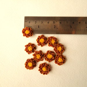 May include: Ten small, brown and yellow artificial flowers, approximately 1 inch in diameter, arranged on a white surface with a ruler for scale.