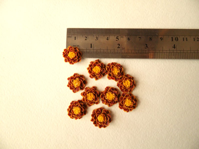 May include: Ten small, brown and yellow artificial flowers, approximately 1 inch in diameter, arranged on a white surface with a ruler for scale.