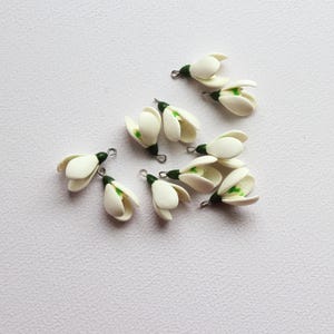 May include: A collection of small, white snowdrop flower charms with green accents and silver-tone findings. The charms are arranged on a white surface, showcasing their delicate, handcrafted details. These floral charms are ideal for jewelry making or craft projects.