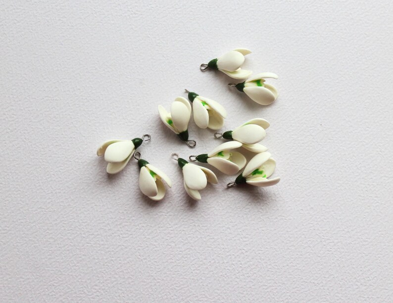 May include: A collection of small, white snowdrop flower charms with green accents and silver-tone findings. The charms are arranged on a white surface, showcasing their delicate, handcrafted details. These floral charms are ideal for jewelry making or craft projects.