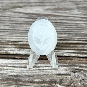 Könnte beinhalten: Ein weißer, ovaler Kristall mit einem geätzten Alien-Gesicht-Design. Der Kristall wird auf einem transparenten Acrylständer vor einer verwitterten Holzoberfläche präsentiert. Die Merkmale des Aliens sind detailliert, mit großen Augen und einem kleinen Mund.