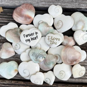 May include: A collection of white, green, and brown heart-shaped stones with engraved messages, including "Forever in my heart", "I Love You", and "7/16/24".