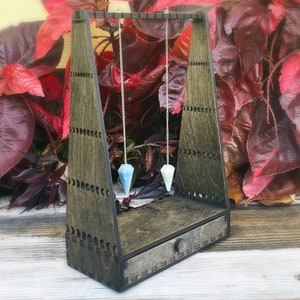 Peut inclure: Un support en bois avec un tiroir pour ranger des pendules. Deux pendules avec des cristaux bleus et verts pendent du support.