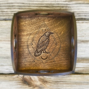 May include: A wooden tray with a dark brown stain and a carved design of a raven inside a circle with geometric shapes. The raven is facing right and has a crescent moon below it.