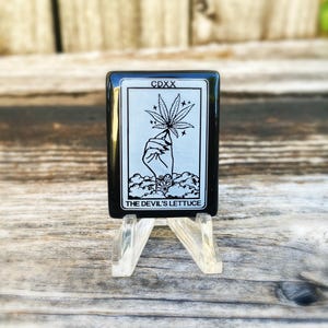 May include: Black and white tarot card with a hand holding a marijuana leaf. The card reads "CDXX THE DEVIL'S LETTUCE".