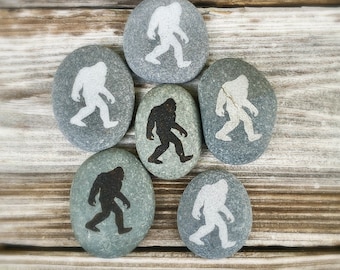 May include: Six gray river stones with Bigfoot silhouettes in black and white. The stones are arranged on a weathered wooden surface. The Bigfoot figures are walking.