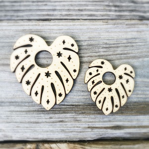 May include: Two wooden heart-shaped ornaments with a cutout design resembling a monstera leaf. The leaves are decorated with black stars and white dots.