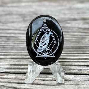 May include: A black, oval-shaped stone with a white raven design inside a geometric pattern. The stone is glossy and has a small hole at the top. It is displayed on a clear acrylic stand.