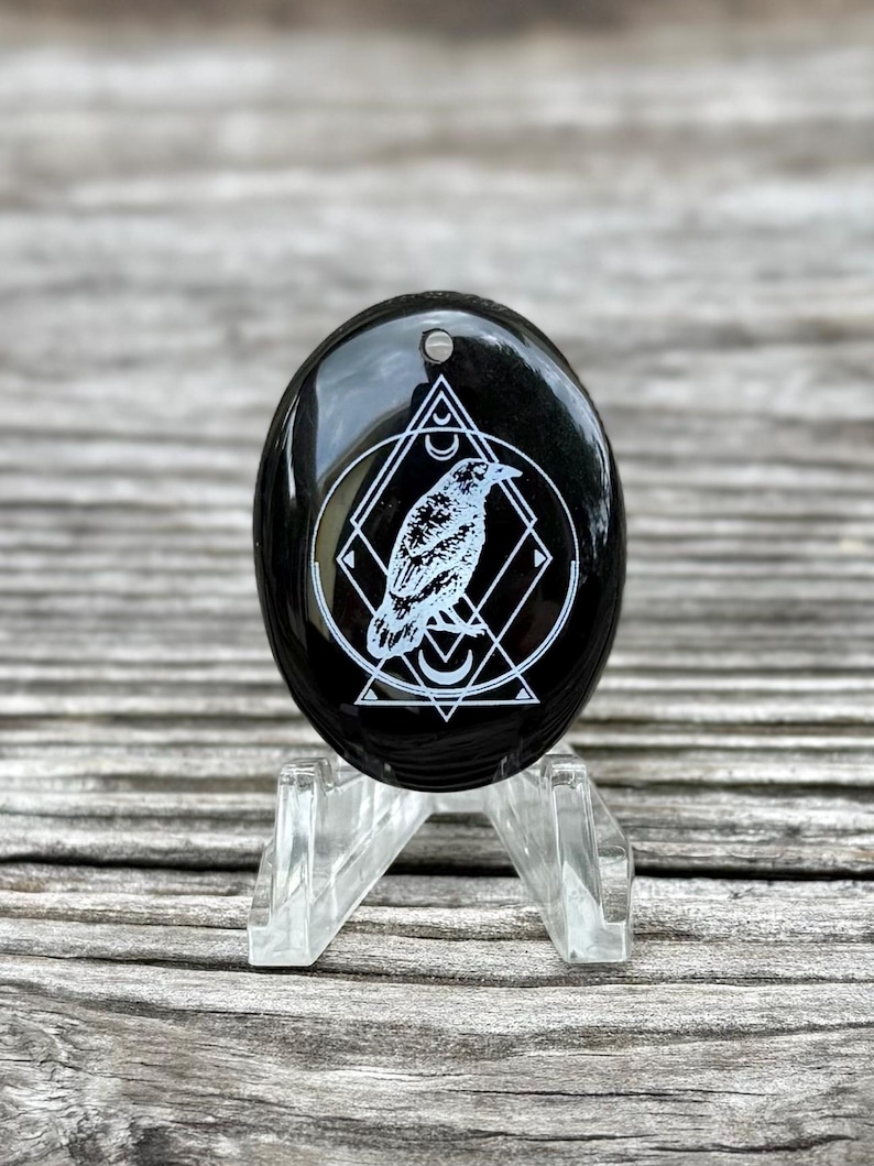 Puede incluir: Una piedra negra de forma ovalada con un dise&ntilde;o de cuervo blanco dentro de un patr&oacute;n geom&eacute;trico. La piedra es brillante y tiene un peque&ntilde;o agujero en la parte superior. Se muestra sobre un soporte de acr&iacute;lico transparente.