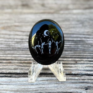 May include: Black obsidian stone with a white etched design of four skeletons dancing under a crescent moon and stars.