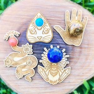May include: Four wooden crystal holders with intricate designs. The holders feature a cat, a snake, a bee, and a hand. Each holder has a different colored crystal in the center.