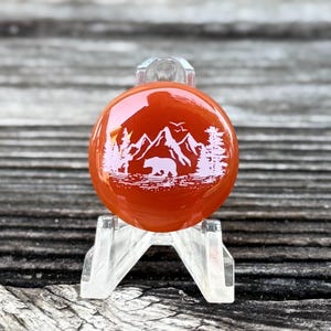 May include: A round, orange glass ornament with a white mountain and bear design. The ornament is displayed on a clear plastic stand, showcasing the detailed landscape scene. The background is a blurred wooden surface.