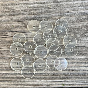 May include: A collection of small, circular, transparent plastic sequins, each featuring a single centered hole. These flat, clear discs are arranged in a loose cluster, showcasing their smooth, reflective surface and uniform round shape.