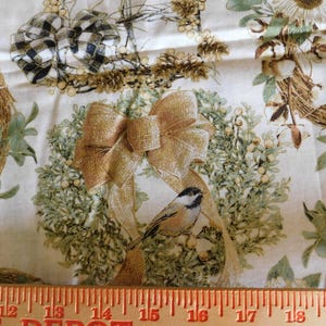 May include: A white fabric featuring a repeating pattern of wreaths decorated with gold and black-and-white plaid bows, sunflowers, and small birds. An orange measuring tape is positioned at the bottom, showing a length of 11 inches.