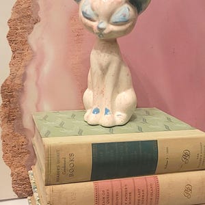 May include: A vintage ceramic cat figurine in a seated position, with a light pink body and blue accents on the ears and eyes. The cat is perched atop a stack of old books with green, pink, and blue spines. The books have titles like "Reader's Digest Condensed Books."