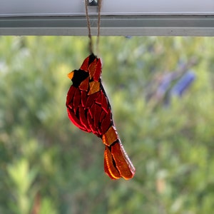 May include: A mosaic cardinal bird ornament, primarily red with gold and black accents, hangs from a natural twine string. The bird's design features a segmented pattern, with a black beak and a detailed tail. The background is a blurred view of green foliage.