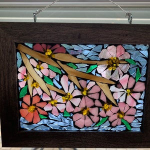Puede incluir: Una obra de arte en mosaico enmarcada que presenta flores rosas y melocot&oacute;n con centros amarillos, hojas verdes y ramas beige sobre un fondo azul. La obra est&aacute; encerrada en un marco de madera marr&oacute;n oscuro, adecuada para la decoraci&oacute;n de paredes.