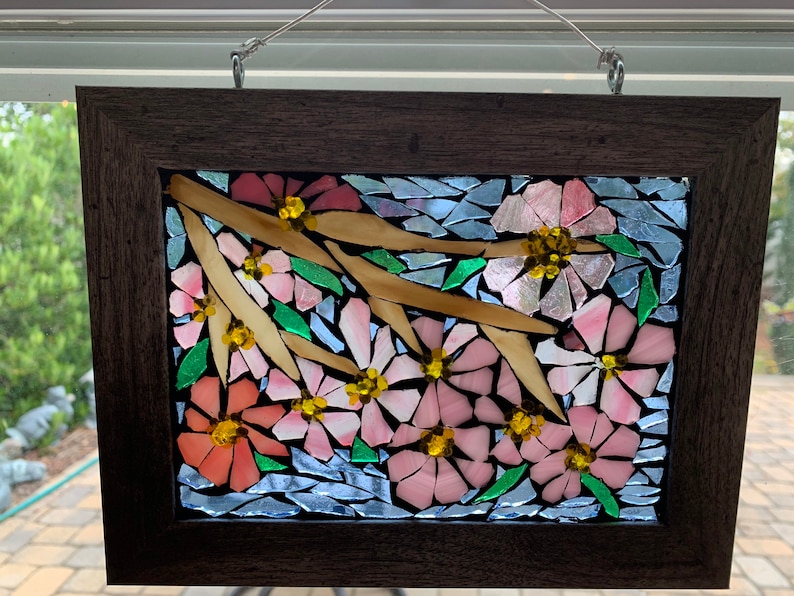 Puede incluir: Una obra de arte en mosaico enmarcada que presenta flores rosas y melocot&oacute;n con centros amarillos, hojas verdes y ramas beige sobre un fondo azul. La obra est&aacute; encerrada en un marco de madera marr&oacute;n oscuro, adecuada para la decoraci&oacute;n de paredes.