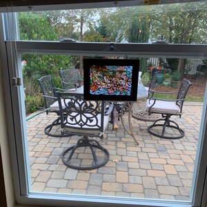 Puede incluir: Una obra de arte en vitral enmarcada que representa flores rosas y naranjas con tallos y hojas verdes. La obra de arte se exhibe frente a una ventana que da a un patio al aire libre con muebles de hierro forjado.