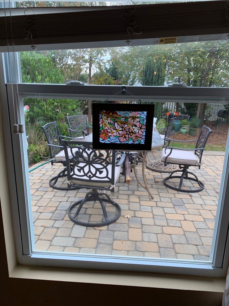 Puede incluir: Una obra de arte en vitral enmarcada que representa flores rosas y naranjas con tallos y hojas verdes. La obra de arte se exhibe frente a una ventana que da a un patio al aire libre con muebles de hierro forjado.
