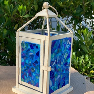May include: A decorative lantern with a white frame and a mosaic of blue glass pieces. The lantern has a hinged door with a glass accent and a metal handle for hanging. The mosaic design features various shades of blue.