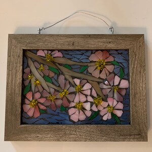 Puede incluir: Una obra de arte en mosaico con flores rosas con centros amarillos y hojas verdes sobre ramas marrones, sobre un fondo azul. La obra est&aacute; enmarcada en un marco rectangular de madera marr&oacute;n claro y tiene un alambre para colgar.
