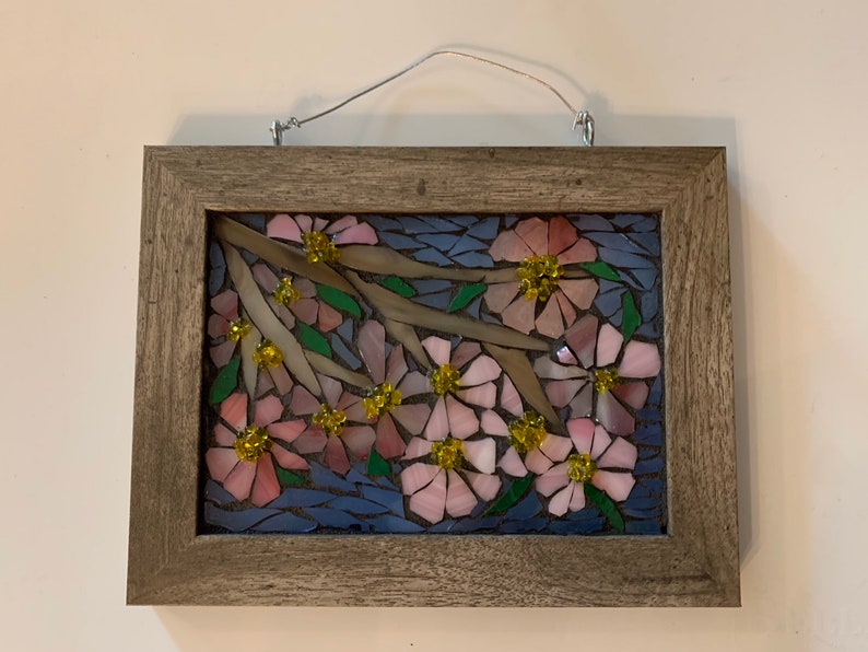Puede incluir: Una obra de arte en mosaico con flores rosas con centros amarillos y hojas verdes sobre ramas marrones, sobre un fondo azul. La obra est&aacute; enmarcada en un marco rectangular de madera marr&oacute;n claro y tiene un alambre para colgar.
