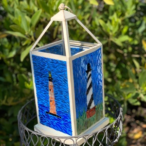 May include: A decorative white metal lantern with blue mosaic panels depicting lighthouses. One lighthouse is orange and the other is black and white. The lantern sits on a decorative metal stand, with a green leafy background.