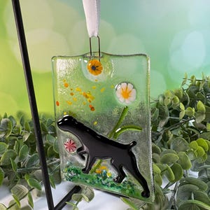 May include: A rectangular glass ornament featuring a black dog silhouette, flowers, and a green background. The ornament is suspended by a white ribbon. The glass has a textured appearance with colorful accents.
