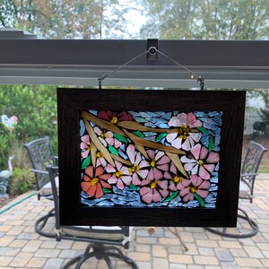 Puede incluir: Una obra de arte en mosaico enmarcada que representa flores rosas y naranjas con centros amarillos y ramas de color marr&oacute;n claro. El mosaico est&aacute; sobre un fondo azul y enmarcado en un marco de madera marr&oacute;n oscuro. La obra de arte est&aacute; colgada frente a una ventana.