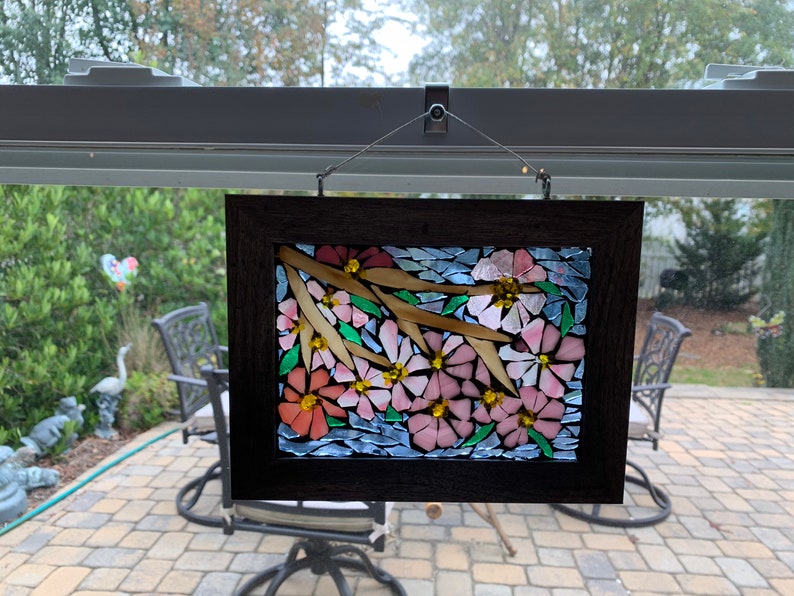 Puede incluir: Una obra de arte en mosaico enmarcada que representa flores rosas y naranjas con centros amarillos y ramas de color marr&oacute;n claro. El mosaico est&aacute; sobre un fondo azul y enmarcado en un marco de madera marr&oacute;n oscuro. La obra de arte est&aacute; colgada frente a una ventana.