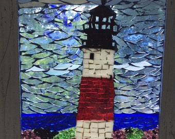 Könnte beinhalten: Mosaik-Kunstwerk, das einen Leuchtturm darstellt. Der Leuchtturm hat einen weißen Turm mit einem roten Abschnitt und einer schwarzen Spitze. Der Himmel ist blau und das Meer ist dunkler blau. Die Basis des Leuchtturms hat grünes und violettes Laub. Das Kunstwerk ist in einem weißen Holzrahmen gerahmt.