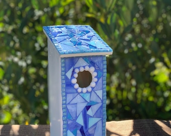 May include: A decorative birdhouse with a mosaic design. The roof and front panel feature a mosaic of blue glass pieces. The birdhouse has a white body and a circular opening with white bead accents. The background is a blurred view of green foliage.