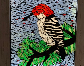 May include: A framed mosaic artwork featuring a woodpecker perched on a branch. The bird has a red head, black and white striped body, and a long black beak. The background is a mosaic of blue and gray pieces, with green leaves.