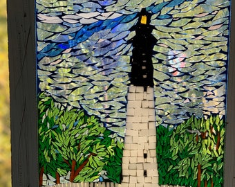 Könnte beinhalten: Mosaikkunstwerk, das einen Leuchtturm mit schwarzem Dach und weißem Turm vor einem bewölkten Himmel zeigt. Grüne Bäume und Gras umgeben den Leuchtturm, der in einem weißen Holzrahmen mit Used-Look eingefasst ist.