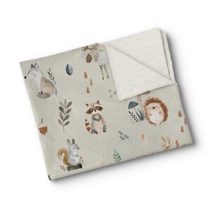 May include: A light green baby blanket with a woodland animal design. The blanket features watercolour illustrations of a fox, raccoon, squirrel, deer, and hedgehog, along with leaves, acorns, and mushrooms. The reverse side is a cream-coloured, textured fabric.