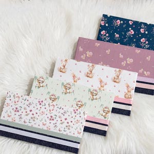 May include: Five rectangular fabric wallets or checkbook covers arranged in a stack. Each features a unique pattern including floral prints, snails, bunnies, and sloths. The bottom edge of each item is accented with horizontal ribbon stripes.