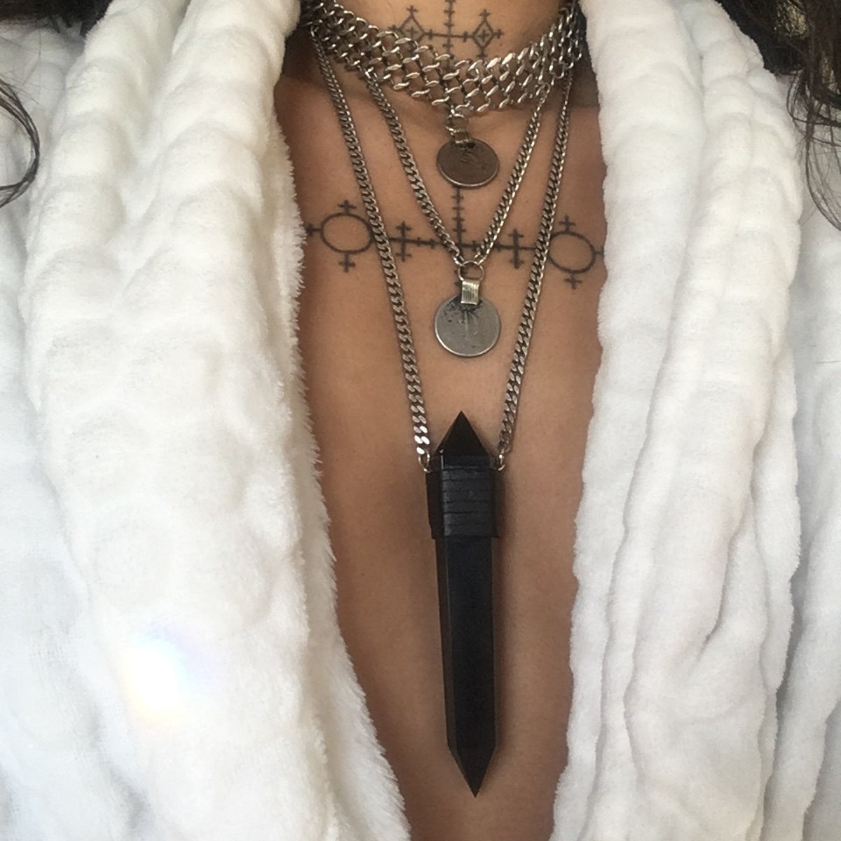 Layered silver coin necklaces with black obsidian pendant worn over chest tattoo and white robe.