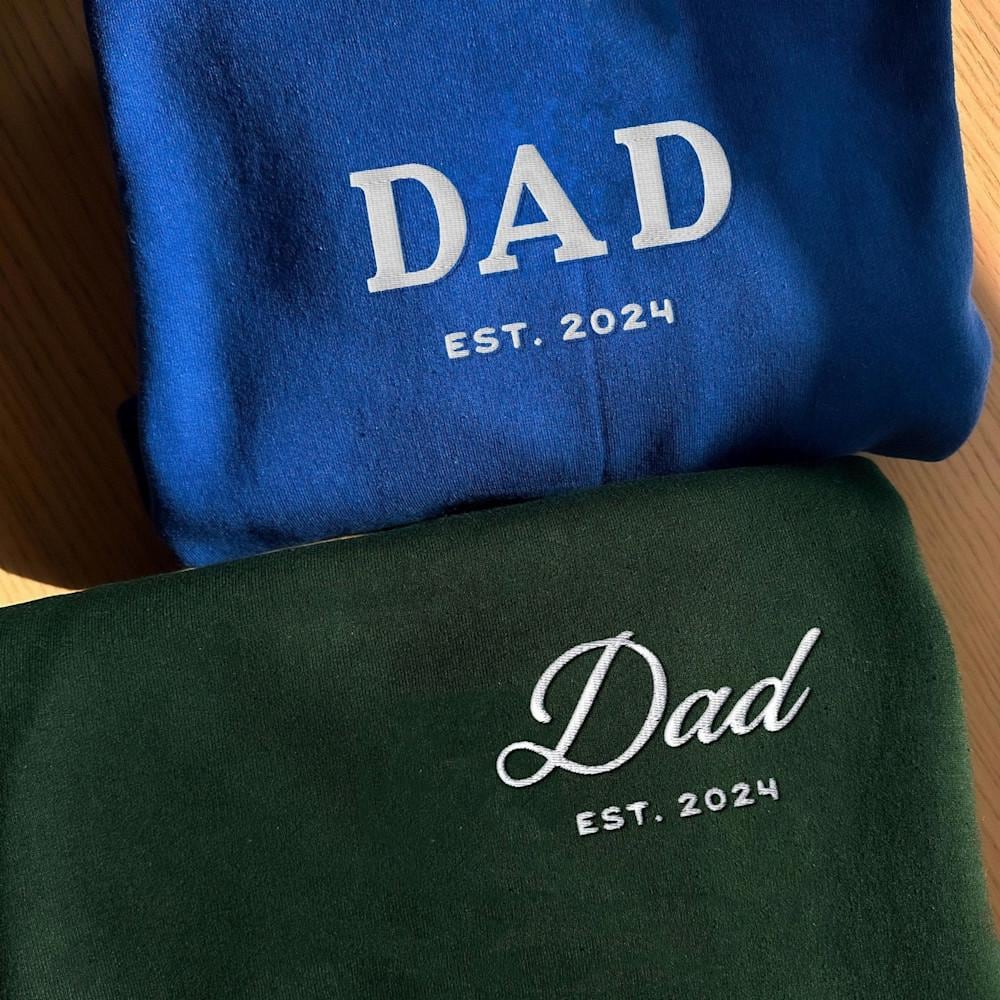 Two custom dad sweatshirts, one green and one blue, embroidered with Dad Est 2024