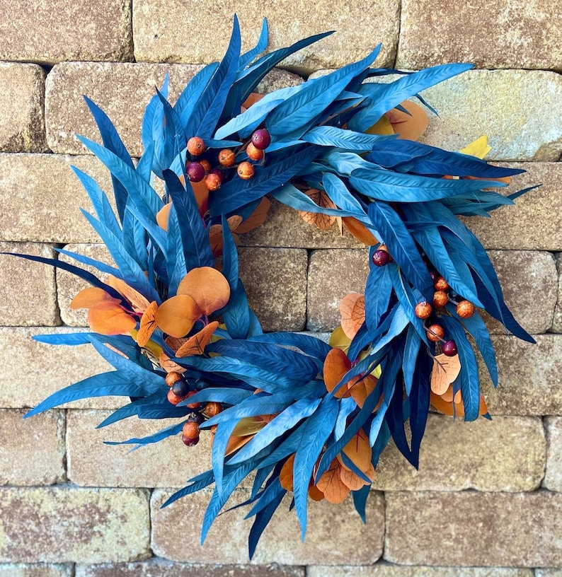 A bright blue and orange fall wreath made from long eucalyptus berries and foliage against a brick background.