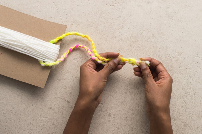 Hands loop top string underneath the tassel body yarn while still attached to chipboard. Hands loop top string underneath the tassel body yarn while still attached to chipboard.