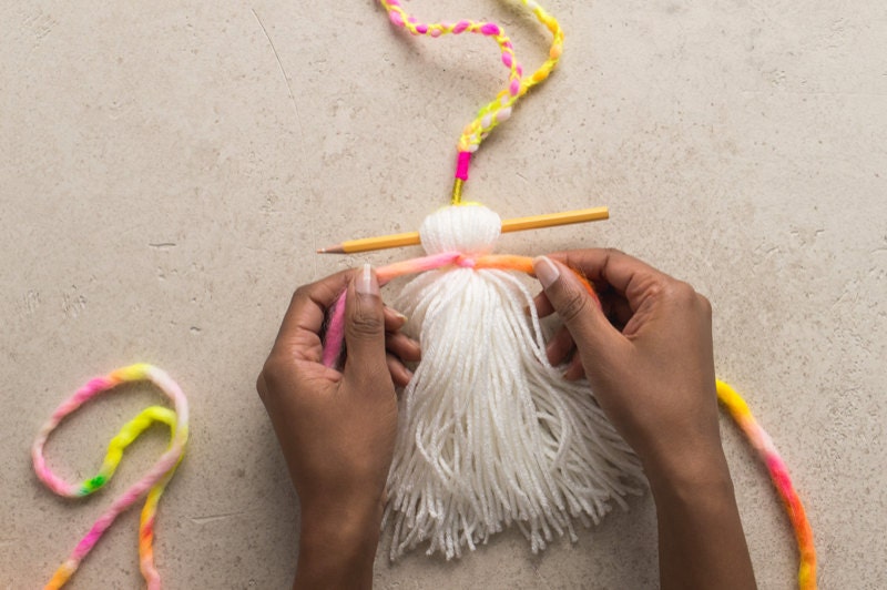 A pencil separates tassel body in two; hands knot yarn around the top of the tassel body. A pencil separates tassel body in two; hands knot yarn around the top of the tassel body.