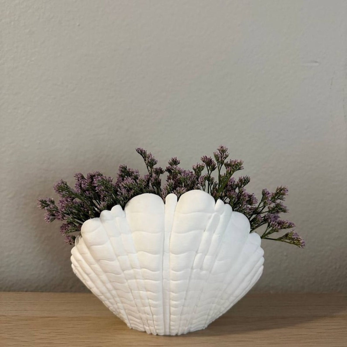 White 3D seashell vase holding purple flowers sitting on a wooden surface.