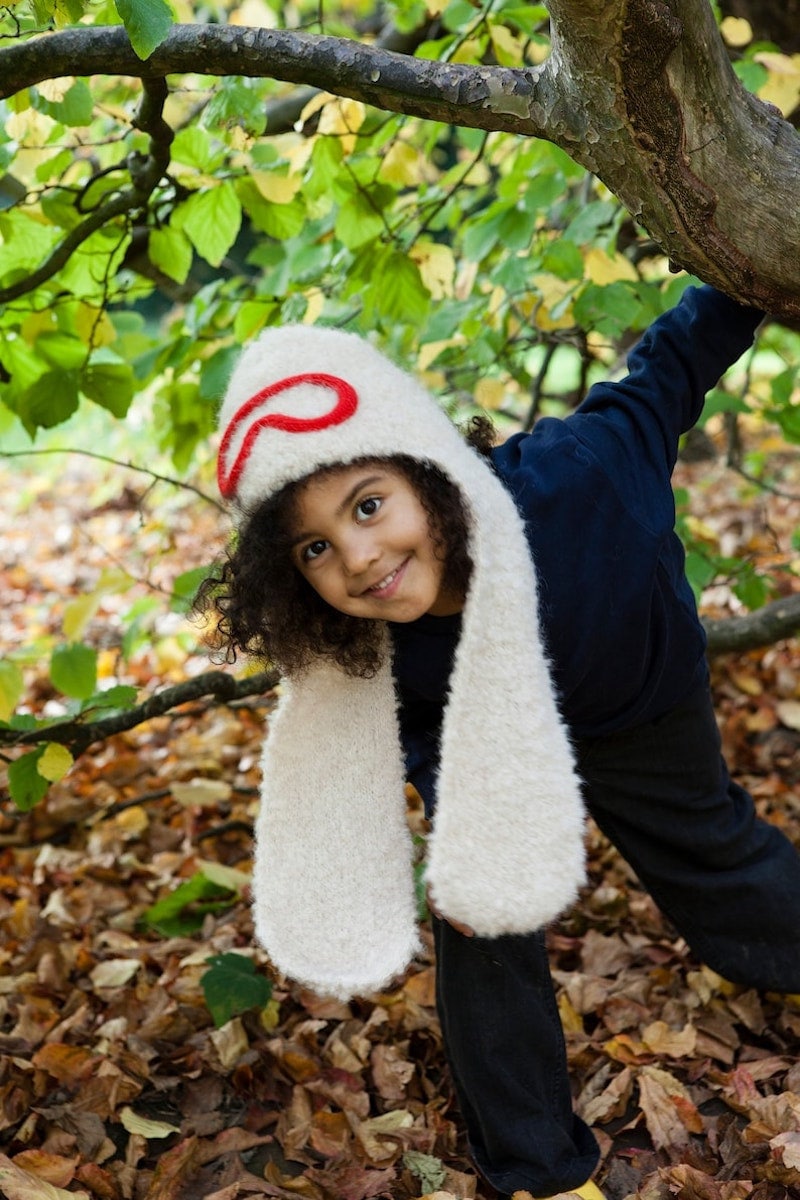 A little girl models a white fuzzy hat with long ear flaps with a red glasses design