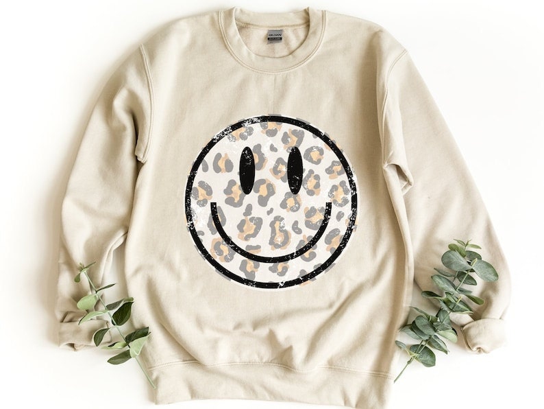 A cream crewneck sweatshirt with a leopard print smiley face on it.