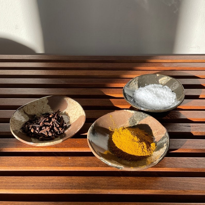 Set of 3 brown and cream handmade ceramic bowls filled with various spices on a wooden table in natural light.